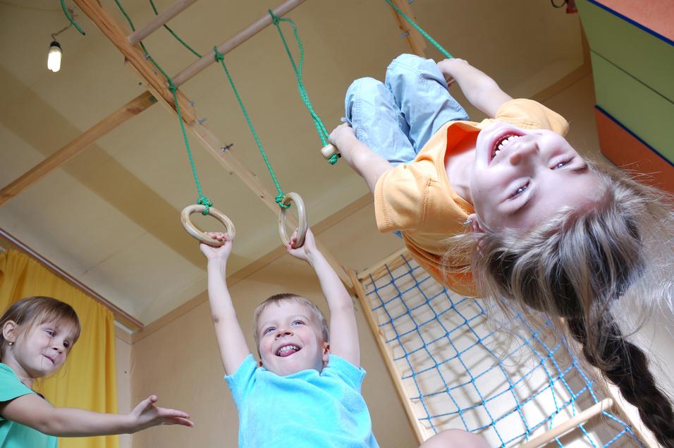 Drei Kinder spielen in einem Raum: Ein Junge hängt an Ringen, während zwei andere lachen und zuschauen.