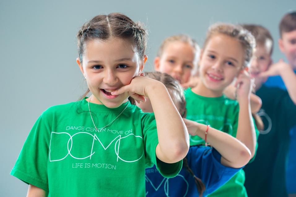 Lächelnde Kinder in bunten T-Shirts machen eine Tanzbewegung vor einem hellen Hintergrund.